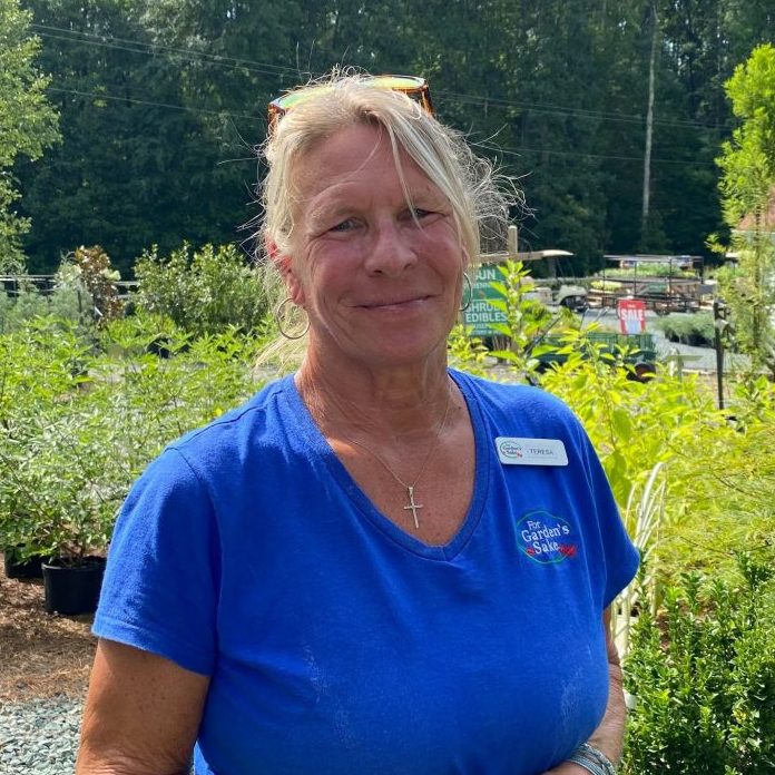 A person wearing a blue shirt and name tag stands in a garden center surrounded by greenery and potted plants, with trees in the background.