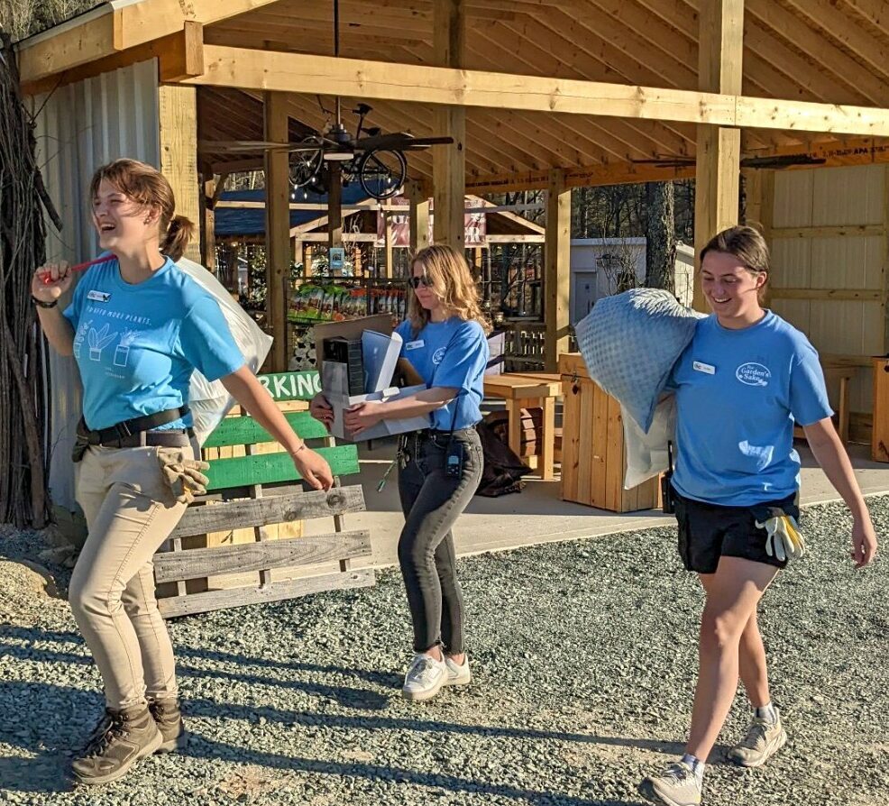 Three people in blue shirts carry supplies near a wooden structure. The sunny outdoor setting suggests a casual work or volunteer activity.