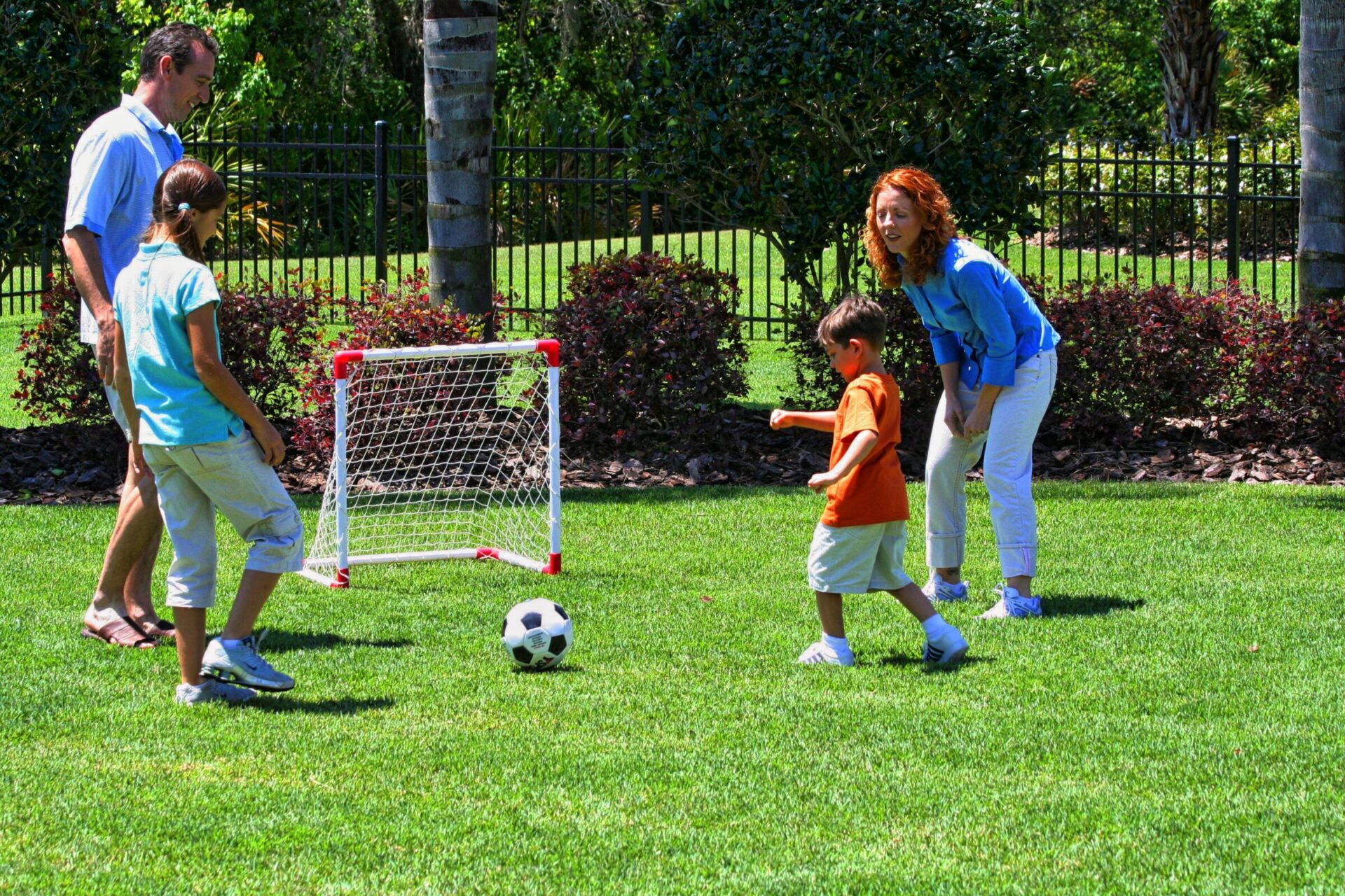 Four people play soccer in a lush garden, enjoying the sunny day. A small goalpost is nearby, surrounded by trees and shrubbery.