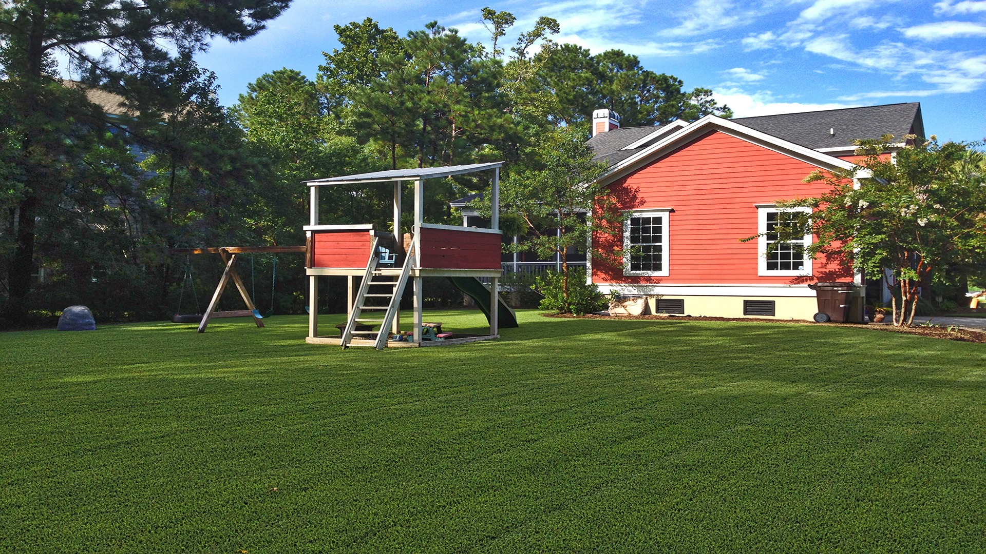 A red house with white trim and a large backyard featuring a wooden playset, surrounded by trees under a blue sky.