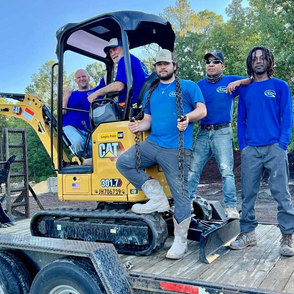 Four people in blue shirts stand around a small excavator on a trailer, surrounded by trees. They appear to be a work crew.