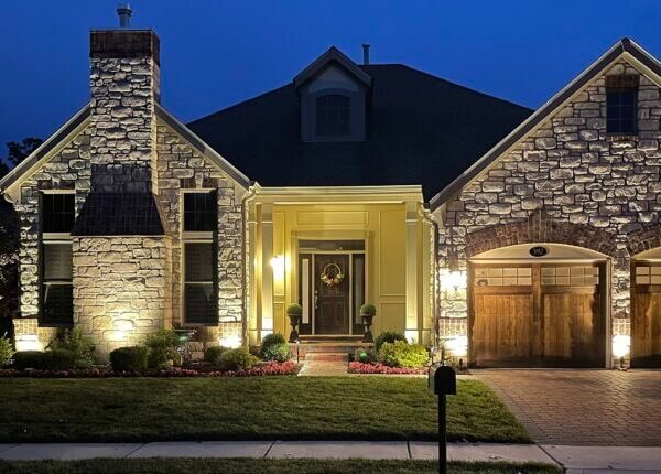 Elegant stone-clad house illuminated by warm accent lighting at night, showcasing the well-maintained front lawn, detailed stonework, and wooden garage doors