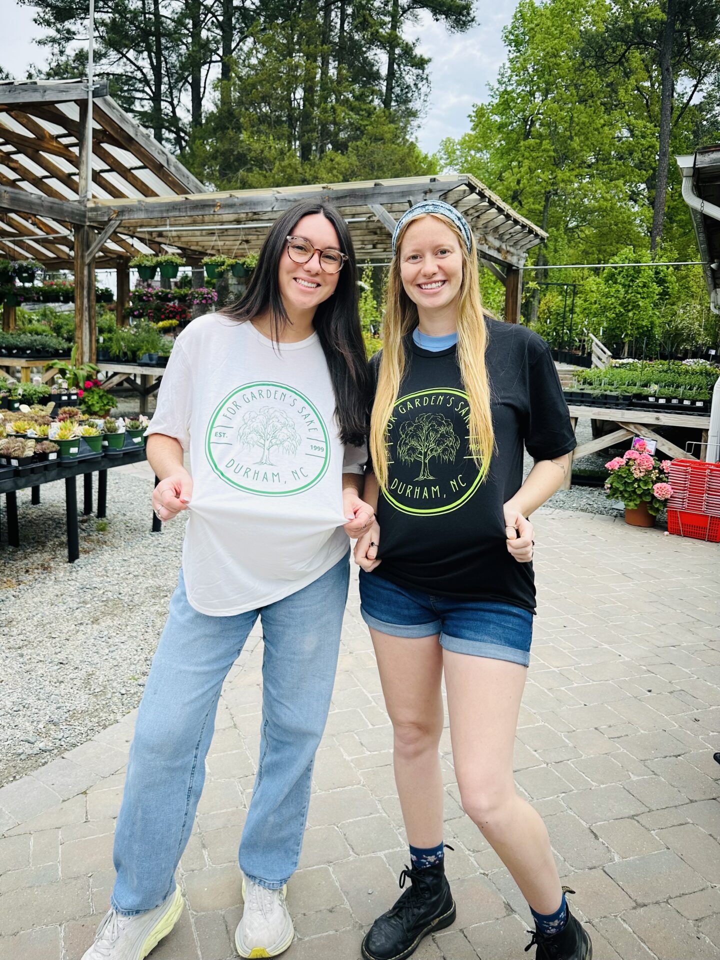 Two people smiling in a garden center, wearing matching t-shirts. Outdoor setting with plants and wooden structures in Durham, NC.