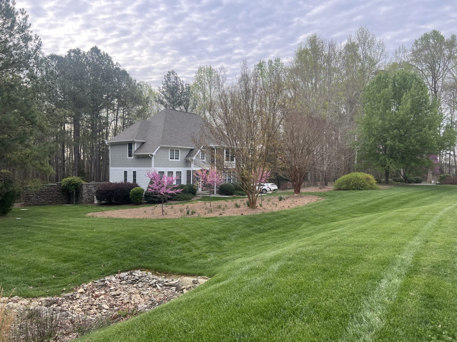 A gray house surrounded by lush trees and a manicured lawn under a cloudy sky, with blooming pink flowers in the garden.