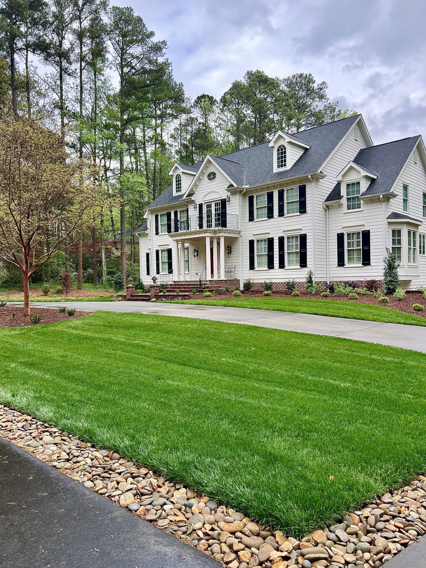 Two-story white house with dark shutters, surrounded by trees. Well-maintained lawn and curved driveway with stone edging. Peaceful suburban setting.