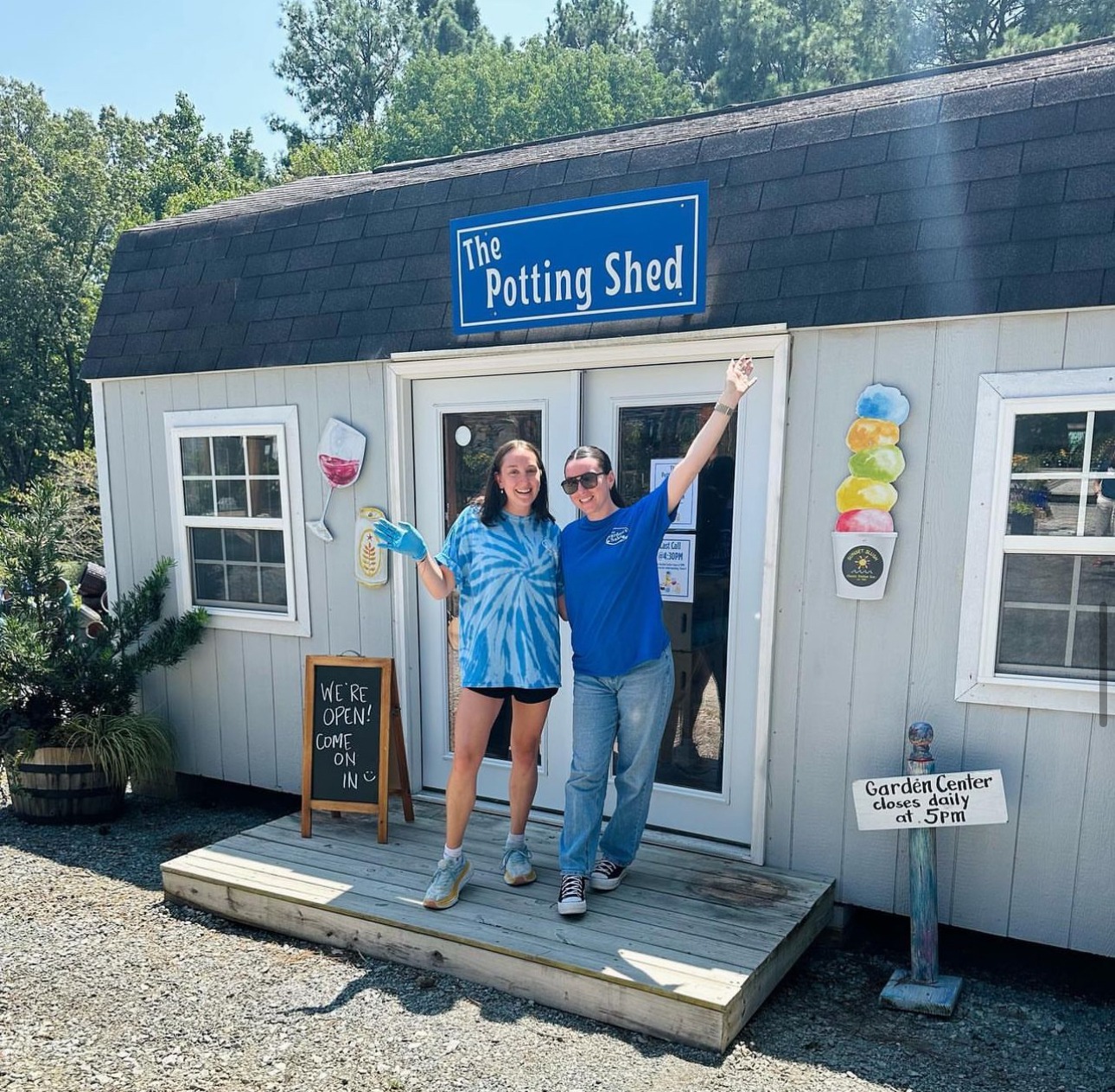 Two people stand smiling outside "The Potting Shed," a small garden shop, with welcoming signs. The weather is sunny, trees in background.