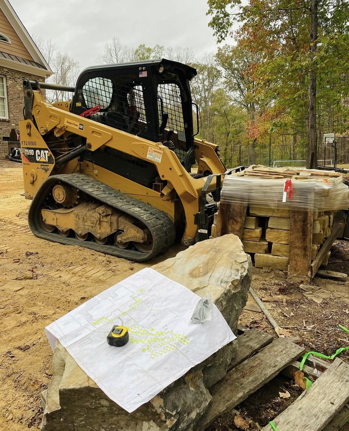 Construction site with a skid steer loader. Blueprint and measuring tape on rock. Stacked stones nearby, surrounded by trees and a building.