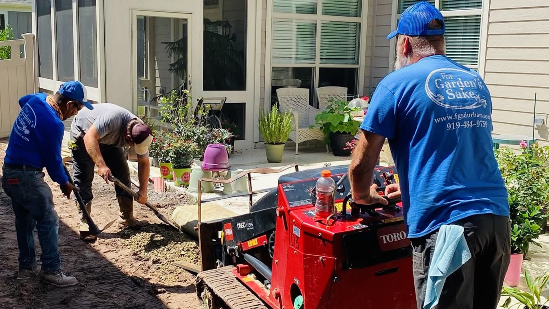 Three people working in a garden, using tools and a small excavator near a house patio. Various potted plants are visible.