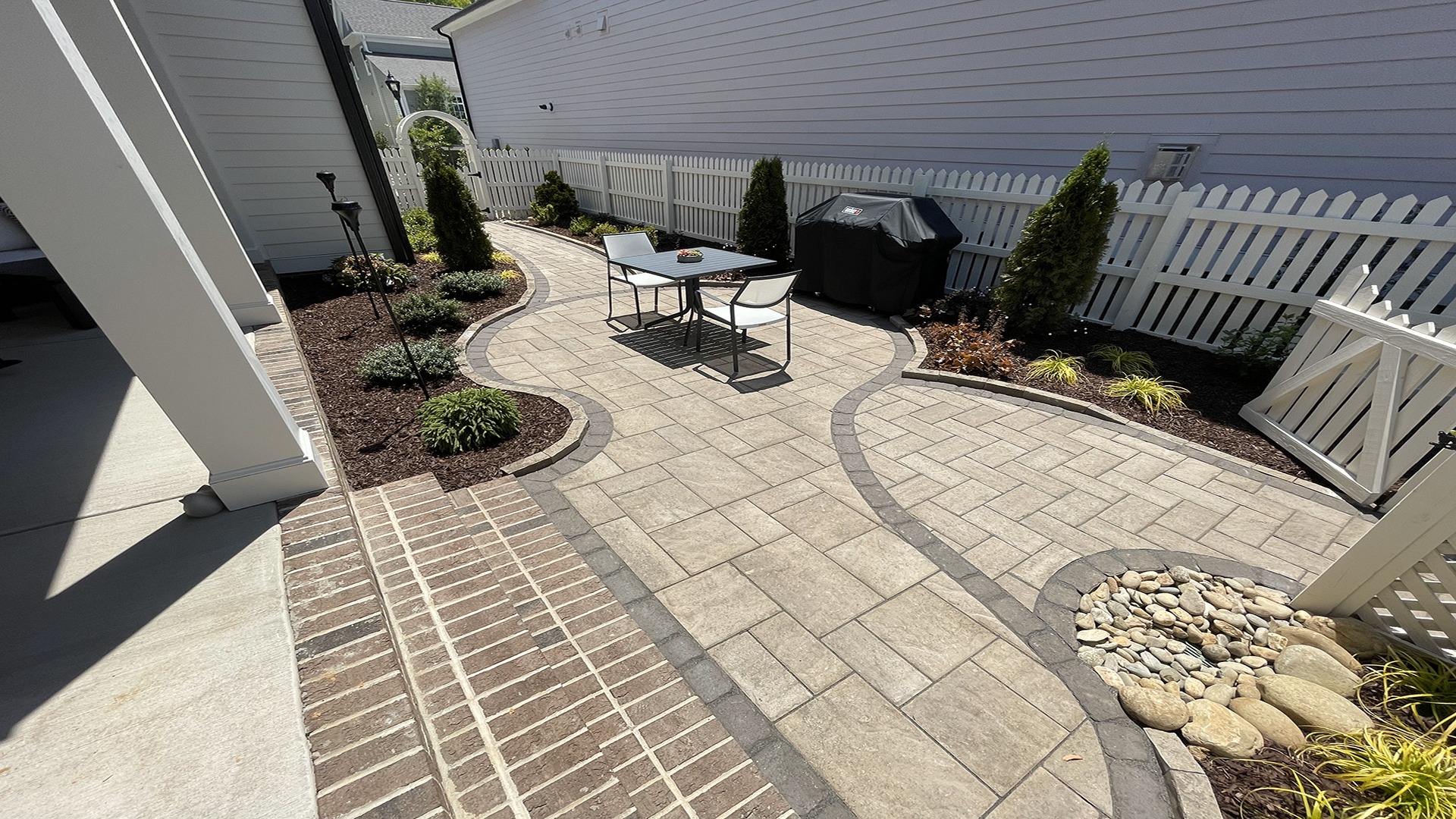 A small, modern backyard features a curved stone path, outdoor dining set, grill, and white picket fence, with neatly arranged plants and mulch.