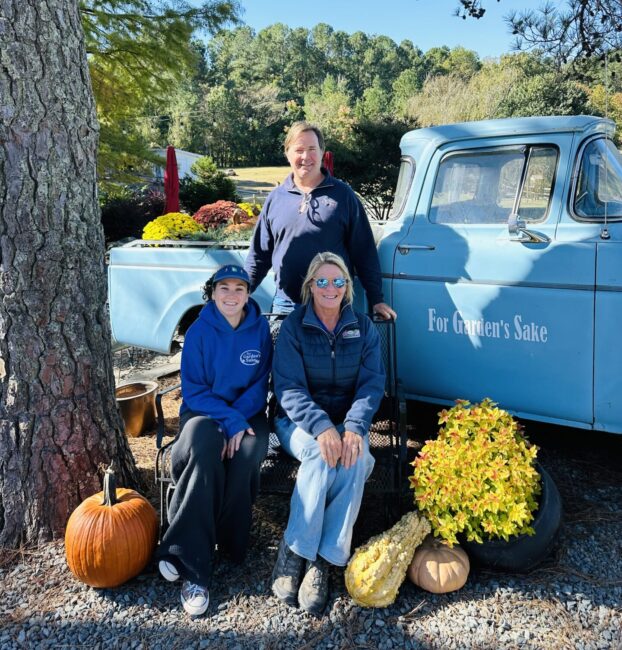 Three people posing by a vintage blue truck labeled "For Garden's Sake," surrounded by pumpkins and autumn foliage in a sunny outdoor setting.