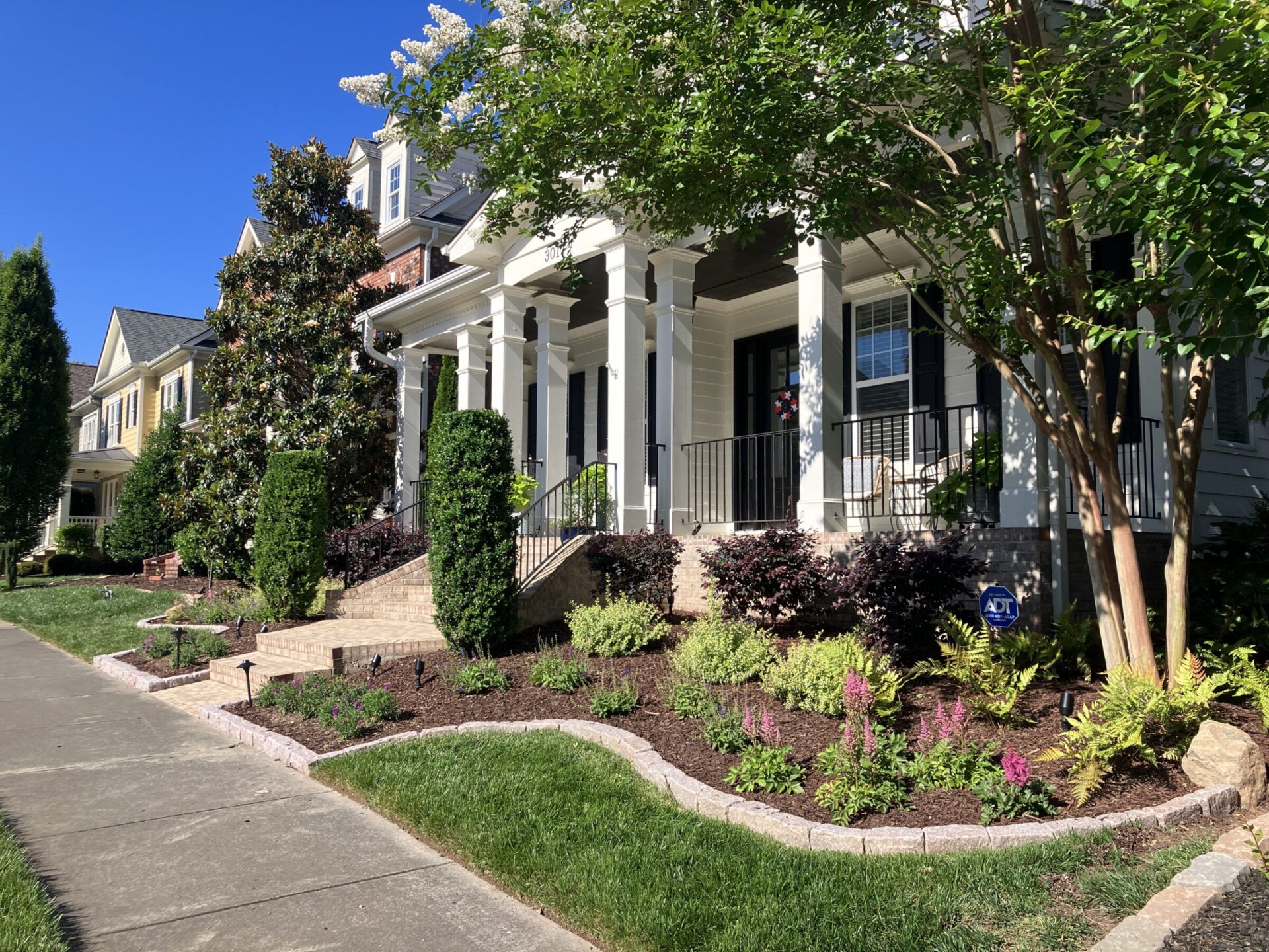 Suburban home with white pillars, landscaped garden, and brick steps. Sidewalk and neatly trimmed lawn under a clear blue sky. Quiet residential street.