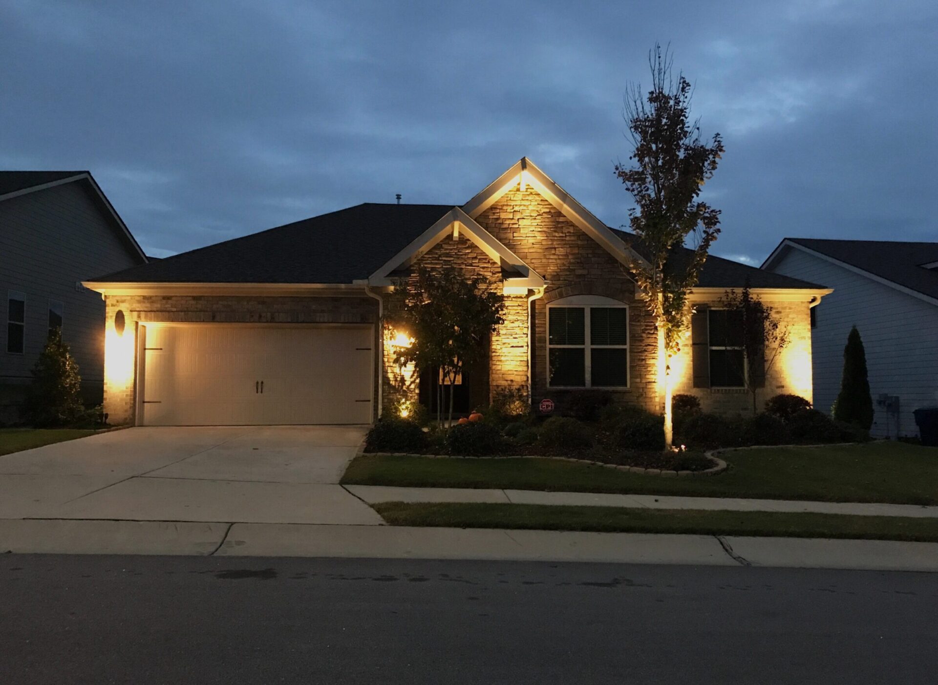 Single-story house with illuminated brick exterior, two-car garage, and landscaped front yard. Evening setting with overcast sky and no visible people.