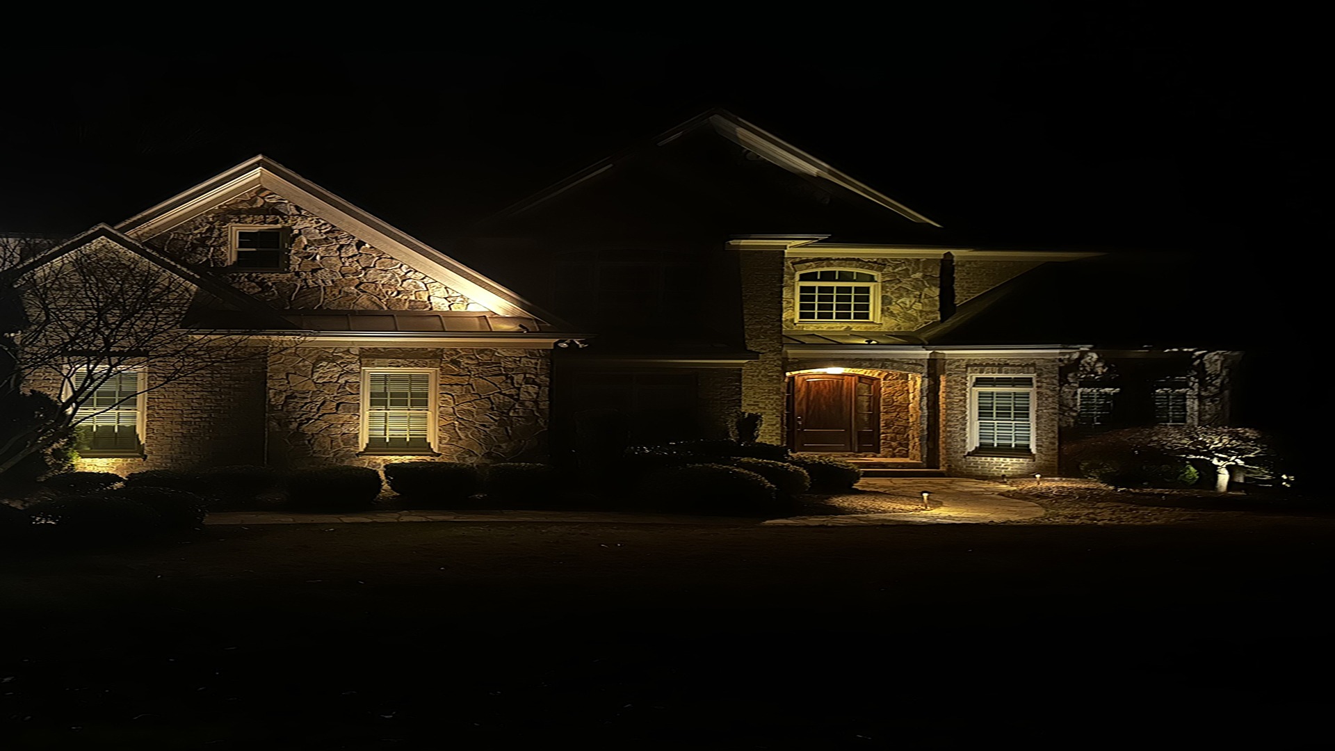 A well-lit, two-story house at night, featuring stone and brick exterior, surrounded by landscaped shrubs and trees, creating a warm and inviting appearance.