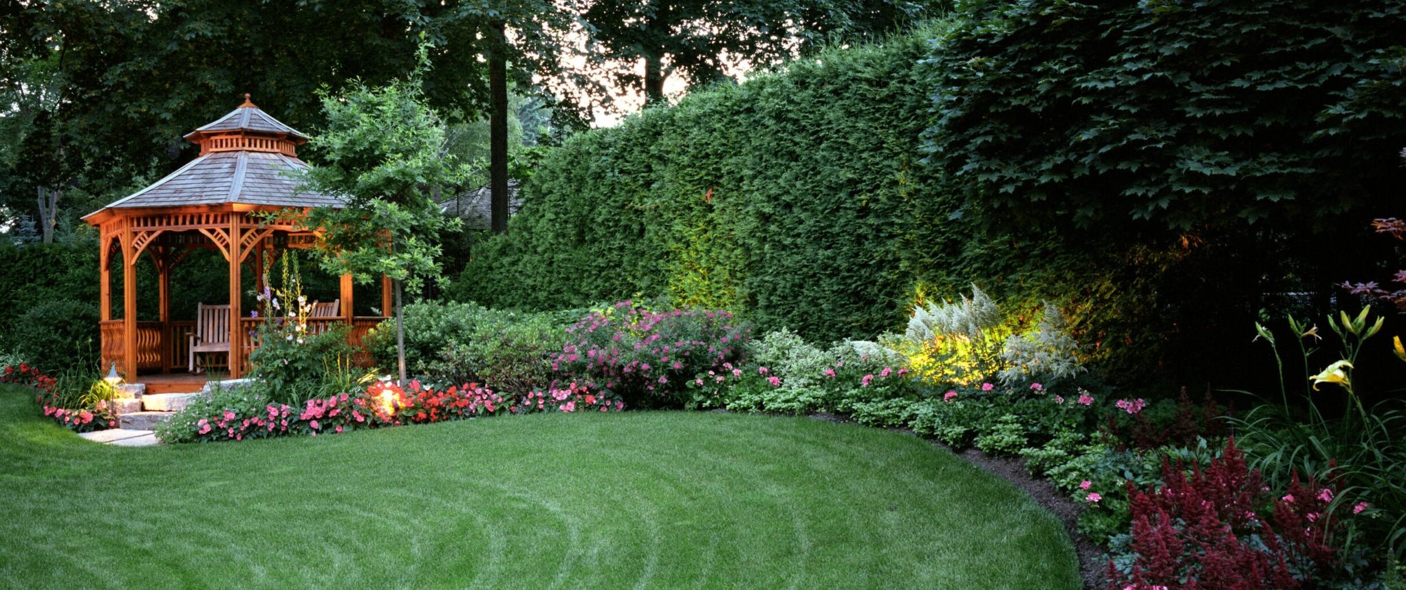 A well-maintained backyard garden with a wooden gazebo, lush green lawn, neatly trimmed hedges, and vibrant flower beds, surrounded by mature trees and landscape lighting at dusk.