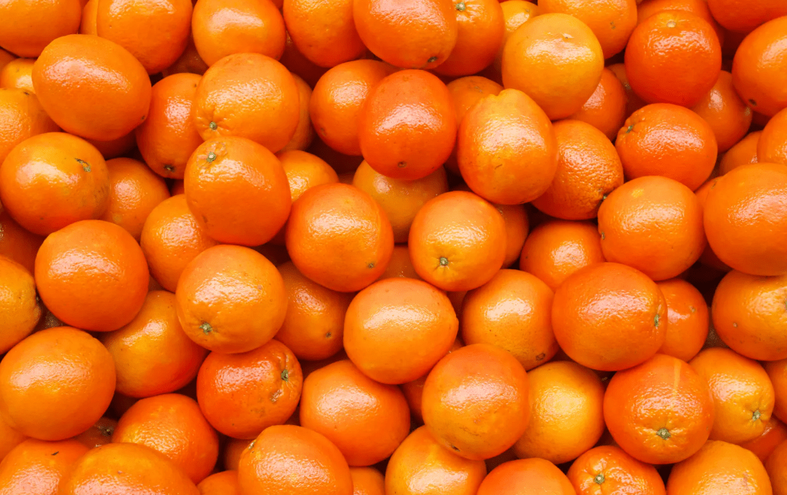 A vibrant pile of fresh, glossy oranges fills the frame, showcasing their vivid color and smooth texture, arranged closely together.