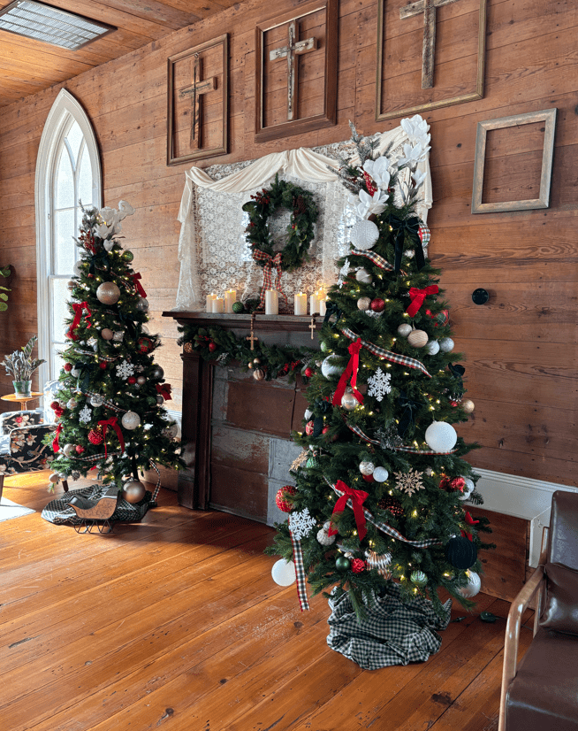 A rustic room with wooden walls, decorated with two Christmas trees, a wreath, candles, and framed crosses above a fireplace.