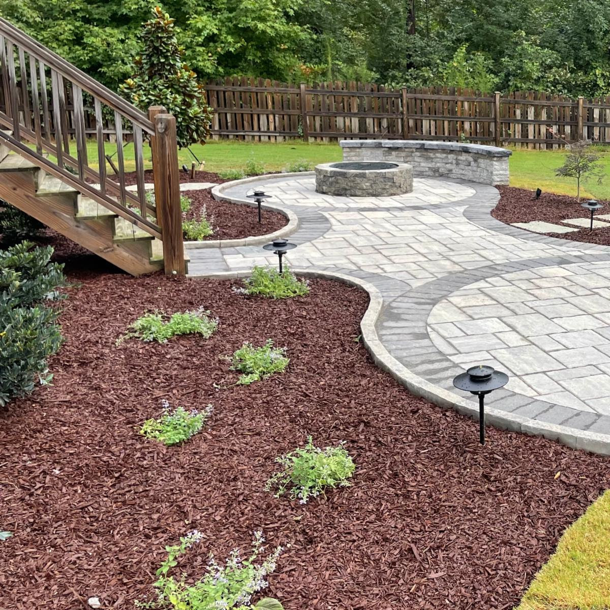 A landscaped backyard features a circular stone patio, wooden stairs, mulch-covered flower beds, ambient lighting, and a wooden fence against lush greenery.