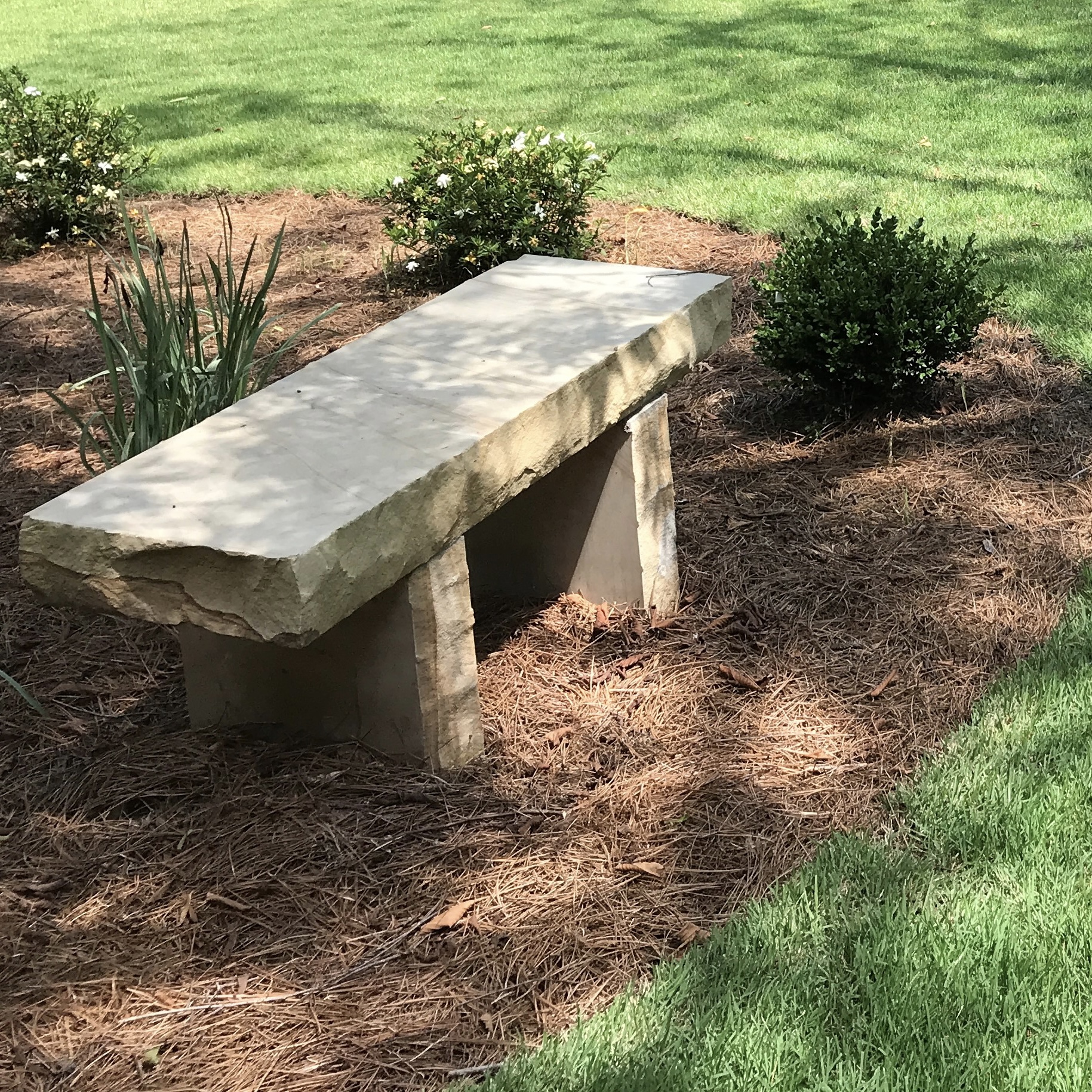 A stone bench is placed on mulch surrounded by neatly trimmed grass and small shrubs, set in a sunny garden area.