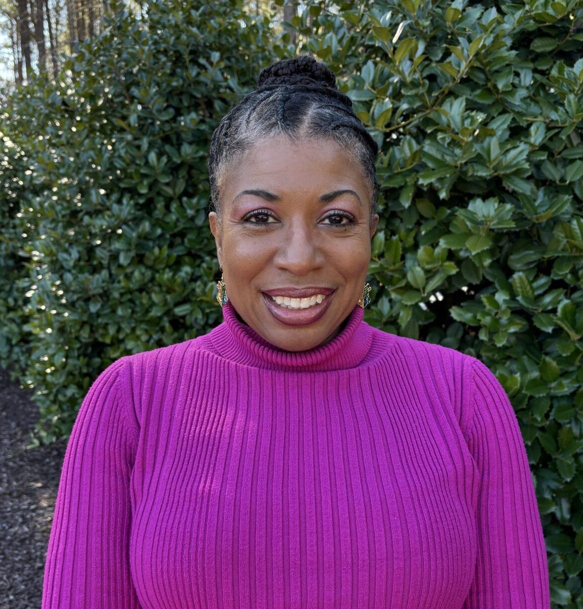 A person in a bright pink sweater stands smiling in front of lush green bushes and tall trees on a sunny day.