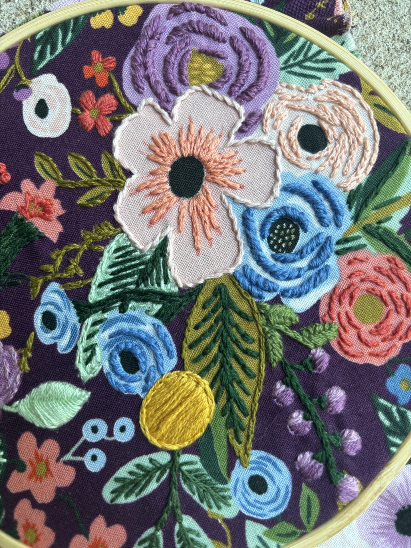 A vibrant embroidery of colorful flowers and leaves on dark fabric, enclosed in a wooden hoop, showcasing detailed stitching.