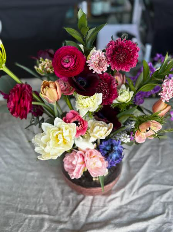 A vibrant bouquet of various colorful flowers arranged in a black vase, placed on a textured gray fabric background.